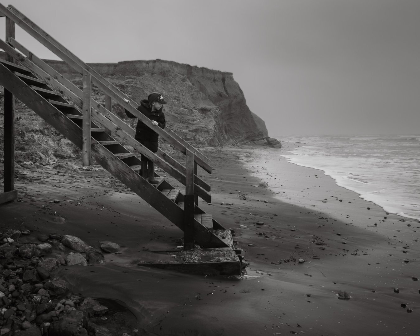Black and white photograph of child on wooden beach steps with eroding cliffs, Isle of Wight — documentary photography by Samuel John Ford