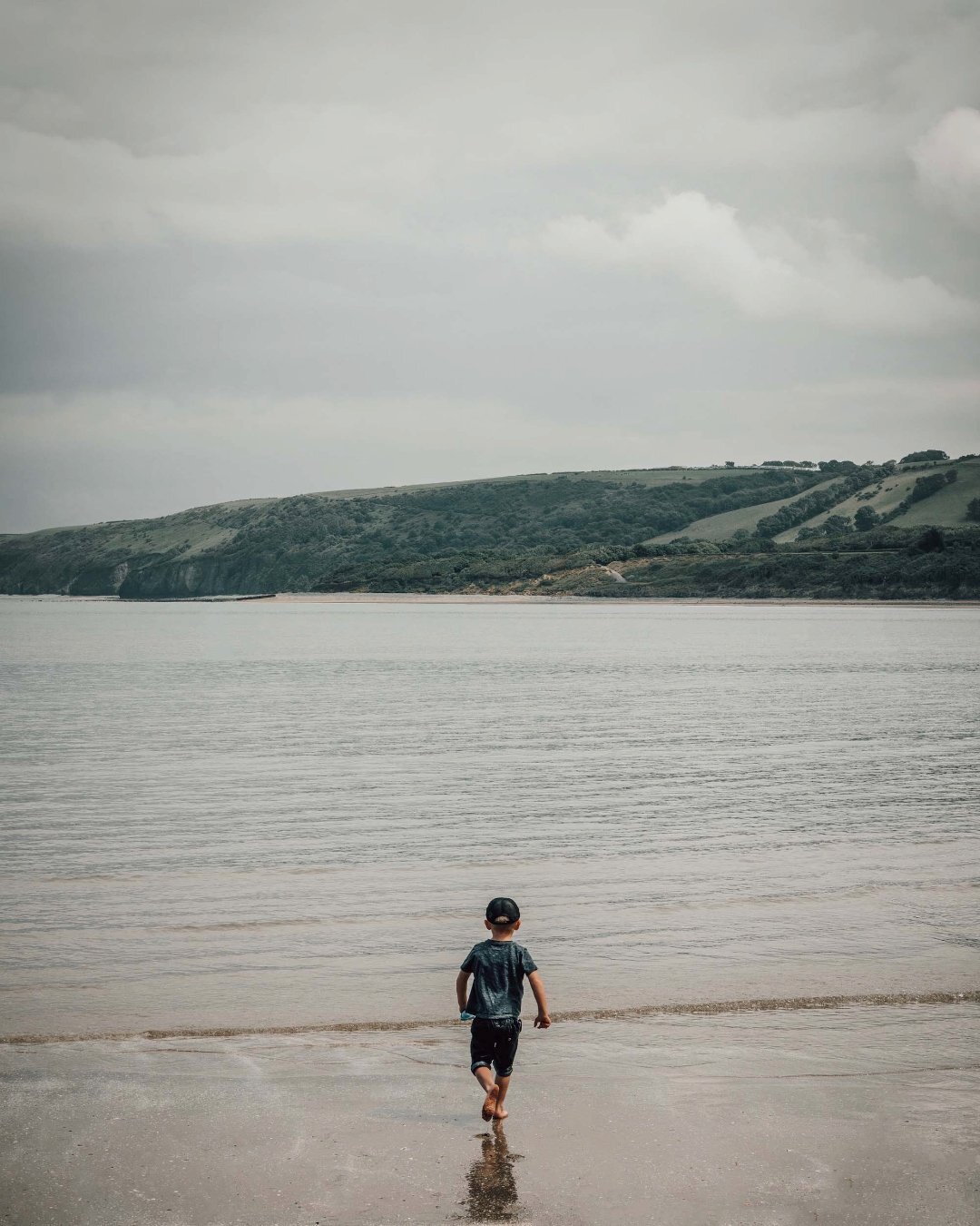 Child walking barefoot into calm sea with green hills behind, Isle of Wight — lifestyle photography by Samuel John Ford