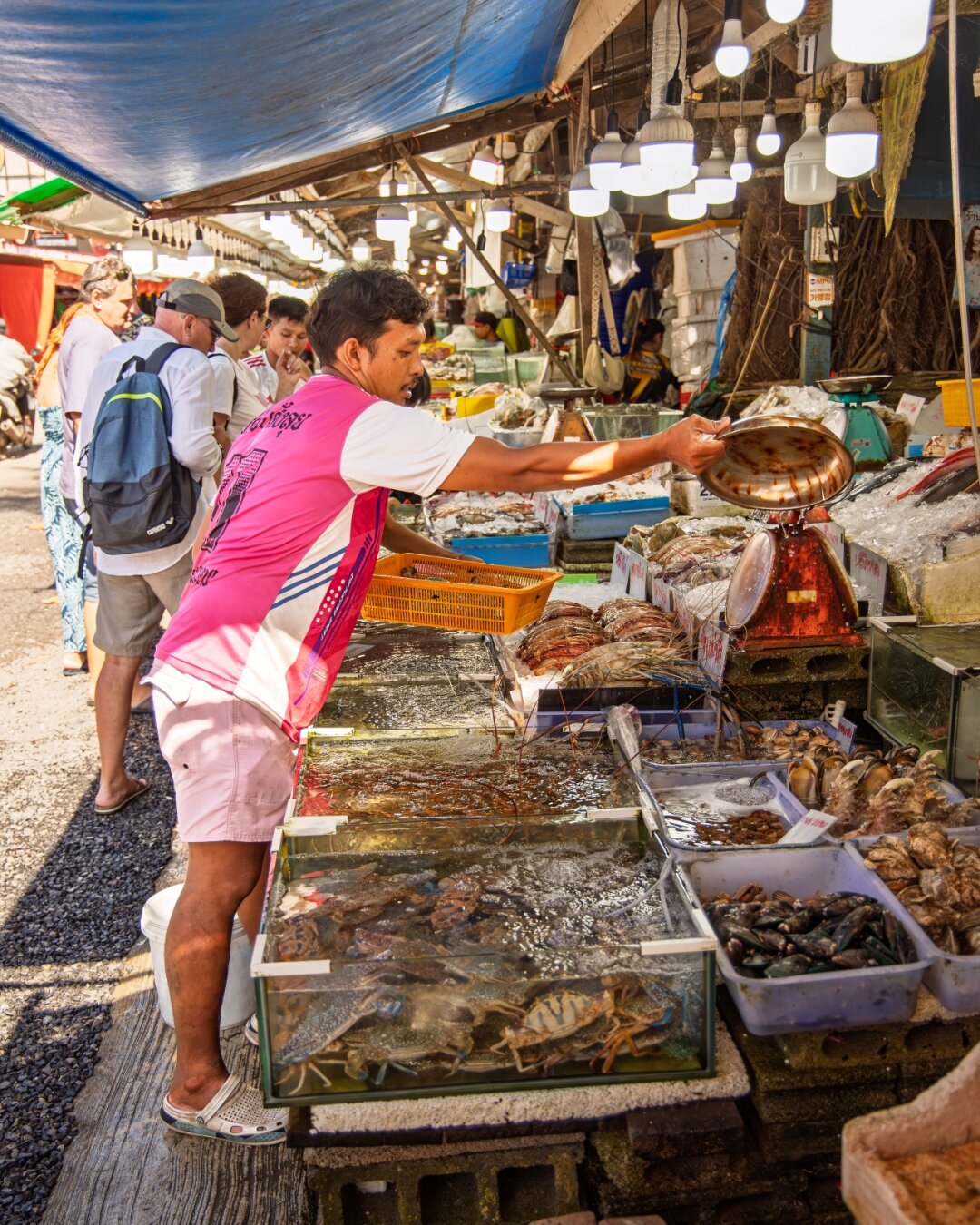 Seafood vendor weighing live prawns at a bustling Thai market stall with tourists browsing — documentary photography